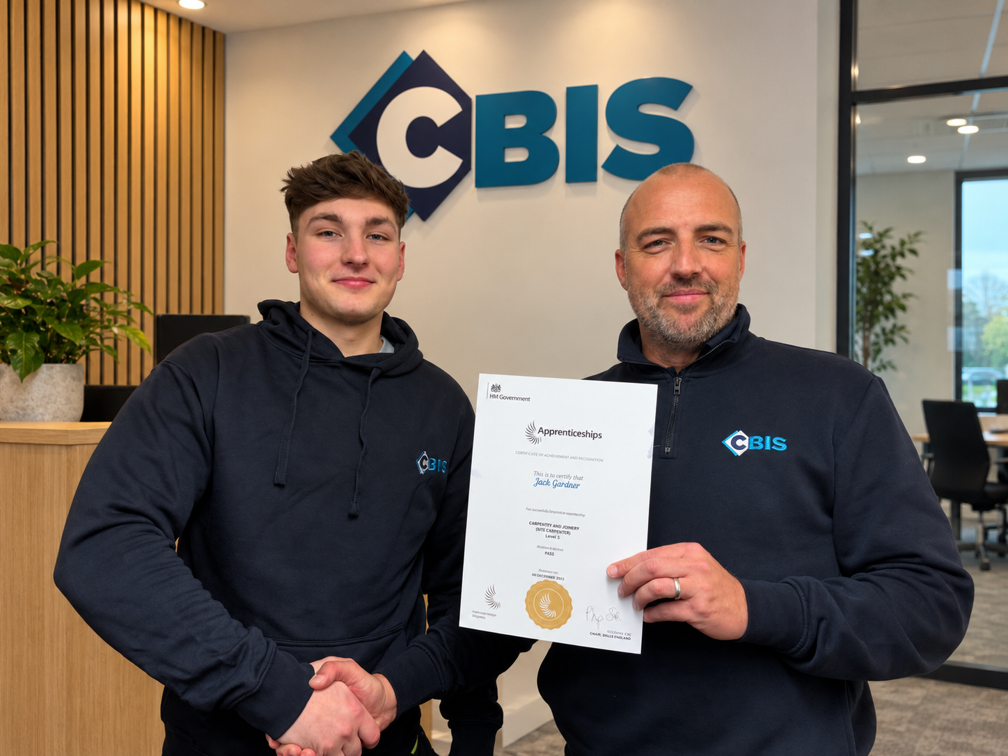 Two people in CBIS workwear holding an apprenticeship achievement certificate in an office with a CBIS wall logo
